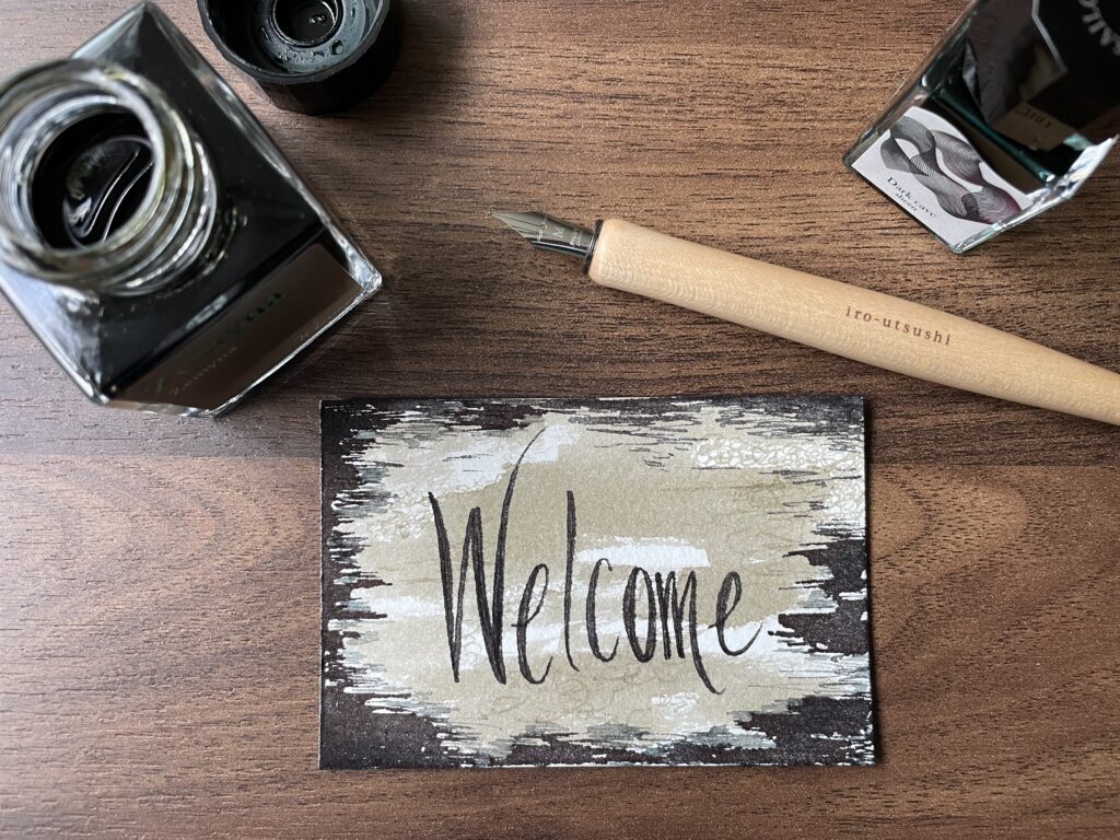 Small artwork with the word 'Welcome' laying on a desk with a dip ink pen and open ink bottle.